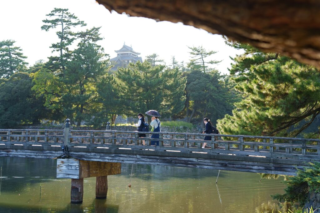 下は松江城の堀川、奥には石垣と松の木、その向こうに松江城の天守閣が見える、宇賀橋という木製の橋と歩く人数人。画像上部にはオープニング同様、松の木の影。