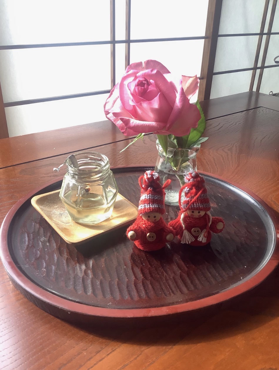 机の上のお盆に、コップに挿された薔薇、毛糸で編まれた人形が置かれている写真。